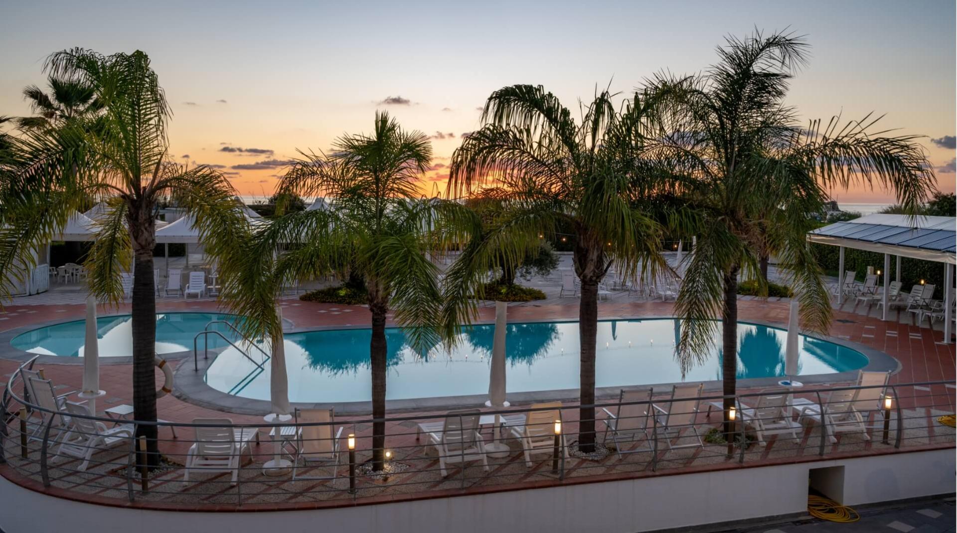 Piscina all'aperto con palme al tramonto, sedie a sdraio e ombrelloni bianchi.
